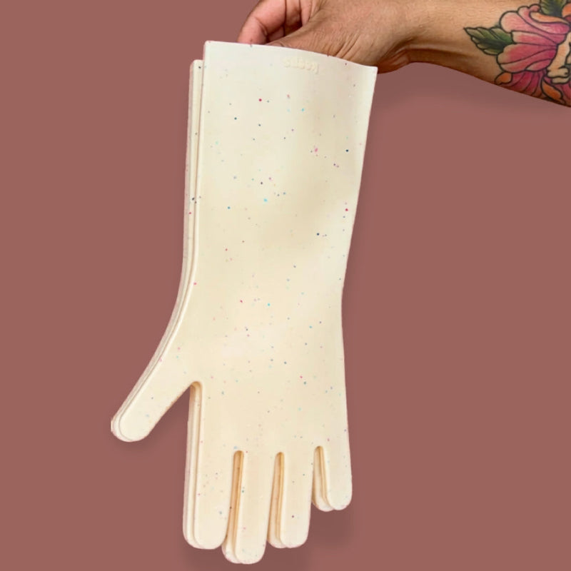 A tattooed hand holds oat speckled sturdy silicone kitchen gloves on a brown background. 