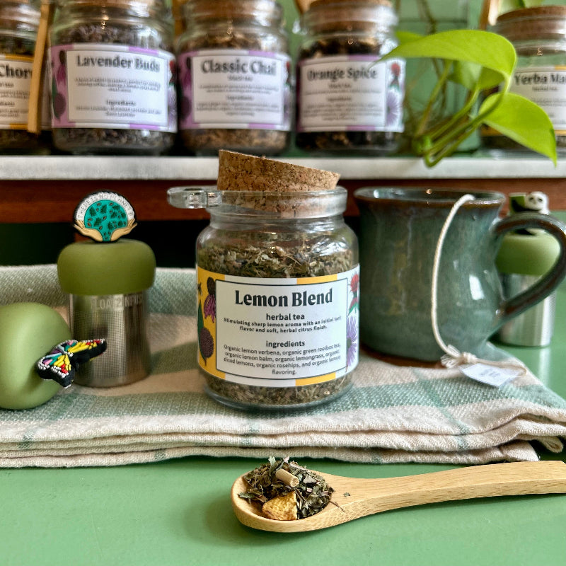 A  refillable jar of lemon blend tea. The jar has a cork lid and a bamboo attachable spoon. The Jar is surrounded by tea steepers and other jars of tea. 