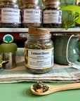 A  refillable jar of lemon blend tea. The jar has a cork lid and a bamboo attachable spoon. The Jar is surrounded by tea steepers and other jars of tea. 