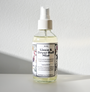 A clear spray bottle of Shade Tree Naturals Linen & Dryer Ball Mist, with floral designs and instructions to add distilled water and essential oils, rests on a white surface in bright natural light against a simple white background.