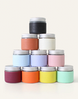 A pyramid of ten pastel or muted Deodorant Cream jars with silver lids by Little Seed Farm, handmade in Tennessee, is arranged on a plain light background.