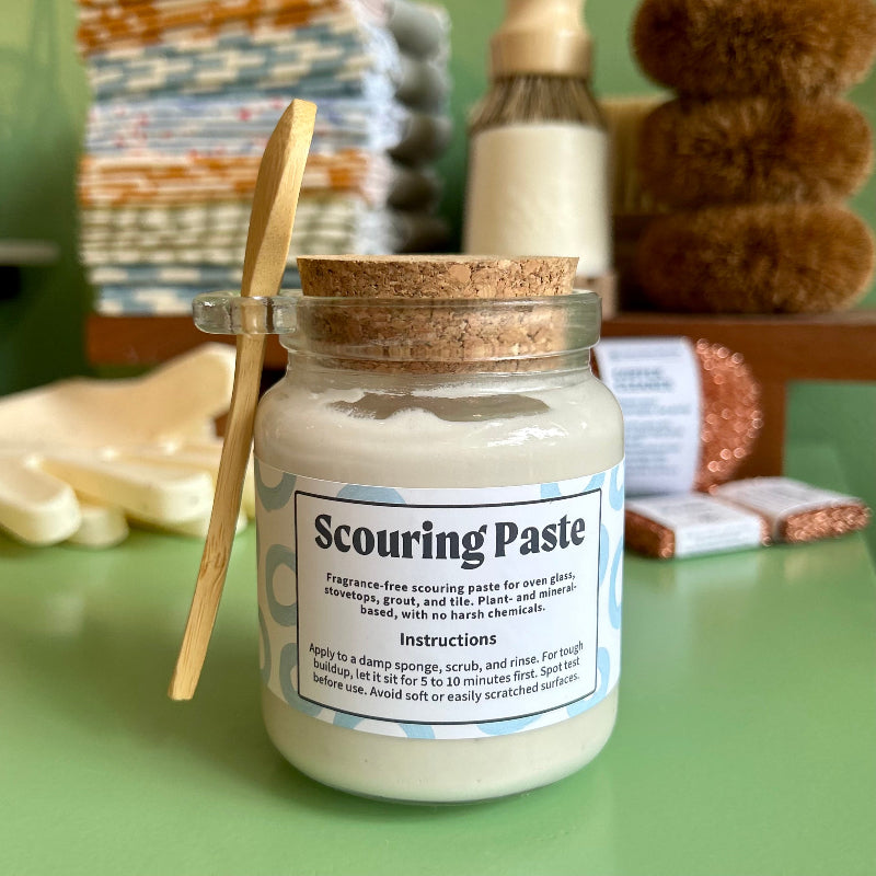 A glass jar with a cork and a bamboo spoon full of plant based scouring paste. The paste is a creamy white and in the background are an assortment of eco-friendly dishwashing supplies. 