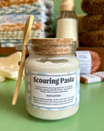 A glass jar with a cork and a bamboo spoon full of plant based scouring paste. The paste is a creamy white and in the background are an assortment of eco-friendly dishwashing supplies. 