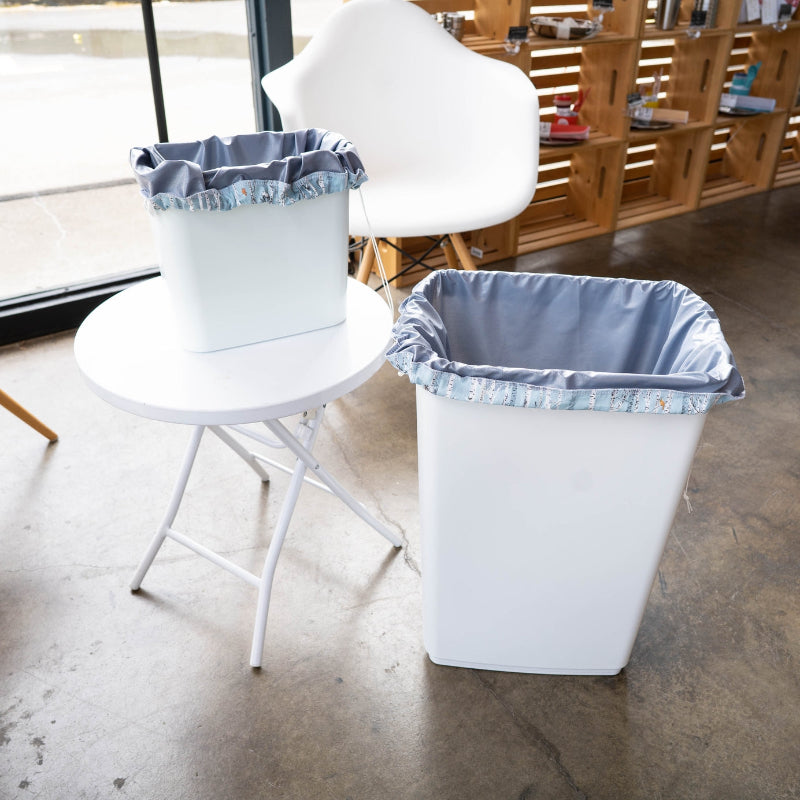 Two white trash bins with grey reusable drawstring pail liners in them. One is a small sized trash bin and the other is a large size. 