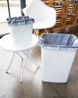 Two white trash bins with grey reusable drawstring pail liners in them. One is a small sized trash bin and the other is a large size. 