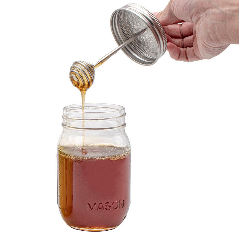 A hand holding the honey dipper mason jar lid as it drizzles honey into a regular mouth mason jar. 
