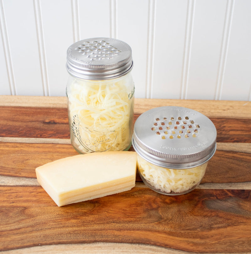 Two mason jars, one regular mouth and one wide mouth each with the stainless steel mason jar grater lid on them. The jars are full of freshly shredded cheese and there is a wedge of cheese next the the jars. 