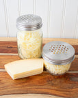 Two mason jars, one regular mouth and one wide mouth each with the stainless steel mason jar grater lid on them. The jars are full of freshly shredded cheese and there is a wedge of cheese next the the jars. 