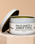 Candle in a tin labeled 'Palo Santo + Amber Topaz' on a beige background