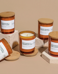 Five Modern Maker Provisions Soy Candles by Modern Makers, each with a wooden lid and unique scent label, are displayed on beige plinths. Hand-poured soy wax promises lasting fragrance; the center candle’s lid is off to reveal the wax inside.