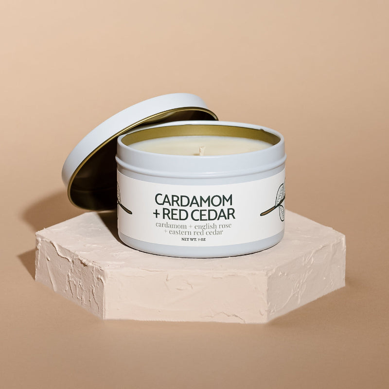 An open white metal candle tin labeled 'CARDAMOM + RED CEDAR' sits on a textured white pedestal against a warm beige background.