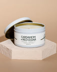 An open white metal candle tin labeled 'CARDAMOM + RED CEDAR' sits on a textured white pedestal against a warm beige background.