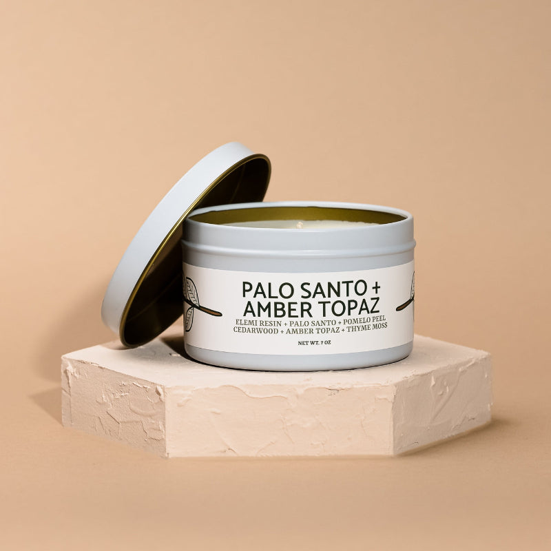 Candle in a tin labeled 'Palo Santo + Amber Topaz' on a beige background