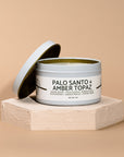 Candle in a tin labeled 'Palo Santo + Amber Topaz' on a beige background