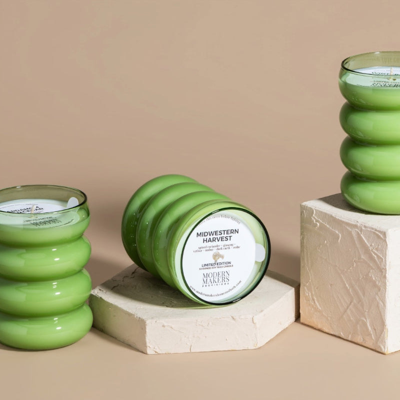 Three Green bubble glass jars with hand poured soy wax candles in them. One in the center is on its side with the label reading Midwestern Harvest facing out. 