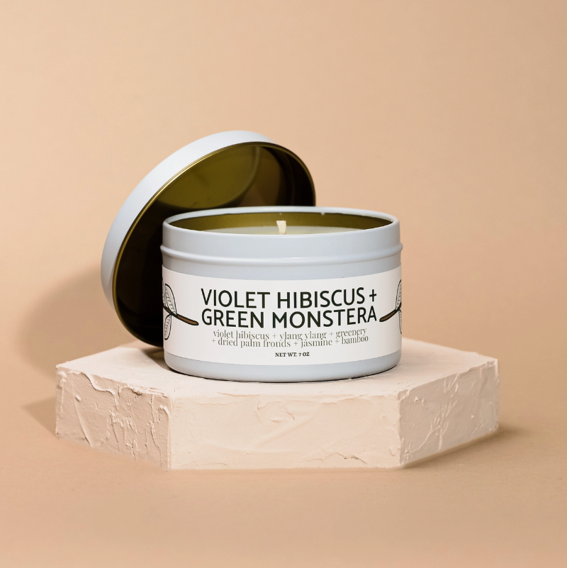 Candle in a tin labeled 'Violet Hibiscus and Green Monstera on a beige background