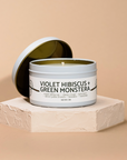 Candle in a tin labeled 'Violet Hibiscus and Green Monstera on a beige background