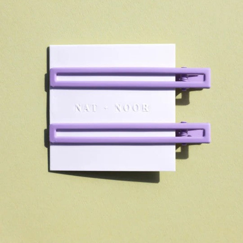 Two thin metal hair clips in light purple. The clips are attached to a white card and are sitting on a green background. 