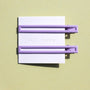 Two thin metal hair clips in light purple. The clips are attached to a white card and are sitting on a green background. 
