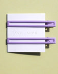 Two thin metal hair clips in light purple. The clips are attached to a white card and are sitting on a green background. 