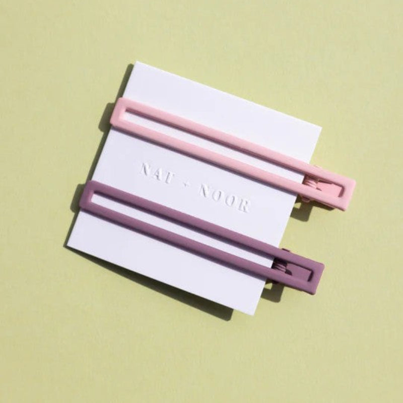 Two thin metal hair clips one is light pink and the other is mauve. The clips are attached to a white card and are sitting on a green background. 