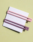 Two thin metal hair clips one is light pink and the other is mauve. The clips are attached to a white card and are sitting on a green background. 