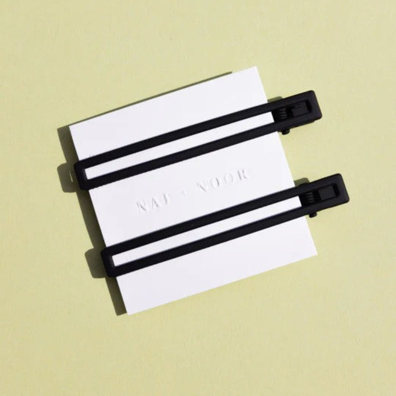 Two thin metal hair clips in black. The clips are attached to a white card and are sitting on a green background. 