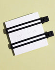 Two thin metal hair clips in black. The clips are attached to a white card and are sitting on a green background. 
