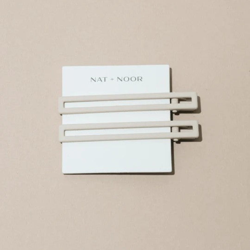 Two thin metal hair clips in white. The clips are attached to a white card and are sitting on a beige background. 