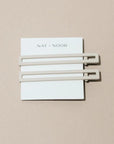 Two thin metal hair clips in white. The clips are attached to a white card and are sitting on a beige background. 