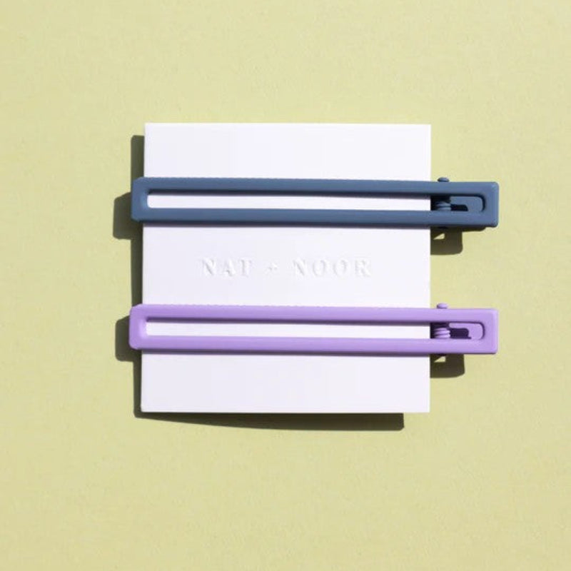 Two thin metal hair clips one is light purple and the other is blue. The clips are attached to a white card and are sitting on a green background. 