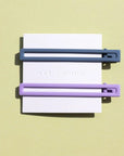 Two thin metal hair clips one is light purple and the other is blue. The clips are attached to a white card and are sitting on a green background. 