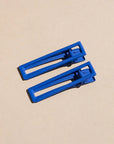 Two wide metal hair clips in bright blue. The clips are sitting on a beige background. 