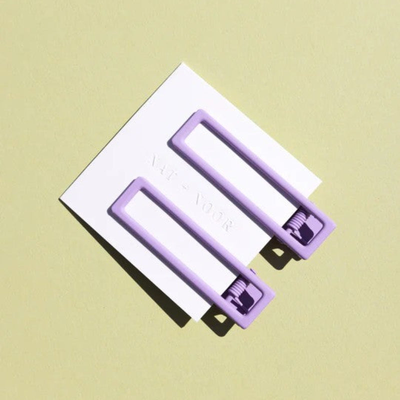 Two wide metal hair clips in a light purple color. The clips are attached to a white card and are sitting on a beige background. 