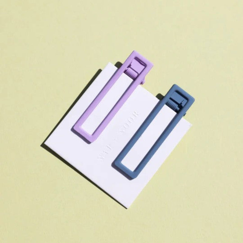 Two wide metal hair clips one is light purple and the other is blue. The clips are attached to a white card and are sitting on a beige background. 