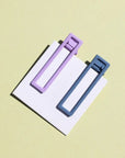 Two wide metal hair clips one is light purple and the other is blue. The clips are attached to a white card and are sitting on a beige background. 