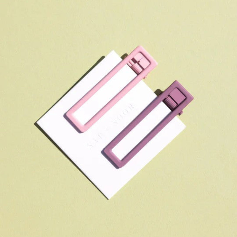 Two wide metal hair clips one is light pink and the other is mauve. The clips are attached to a white card and are sitting on a beige background. 