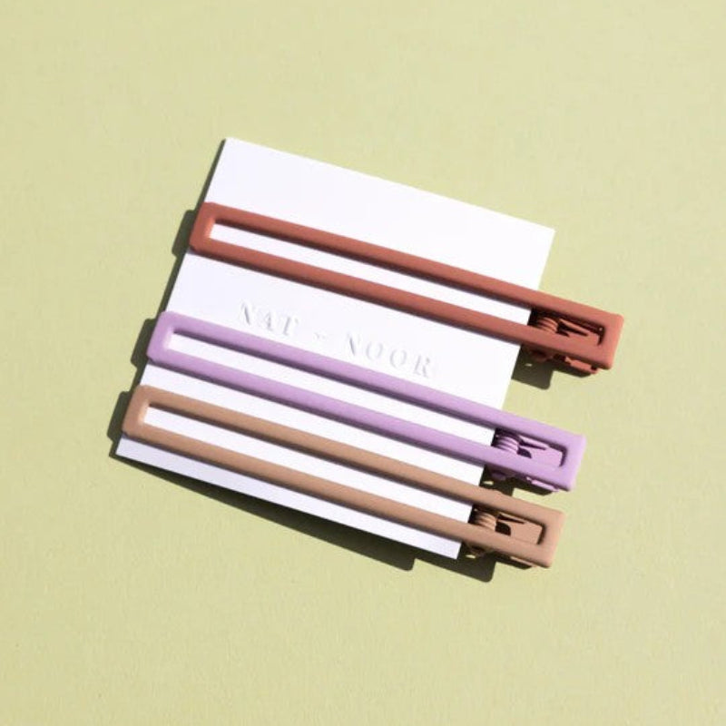 Three metal hair clips in orange, purple, and tan. The clips are attached to a white card and on a green background. 