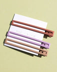 Three metal hair clips in orange, purple, and tan. The clips are attached to a white card and on a green background. 