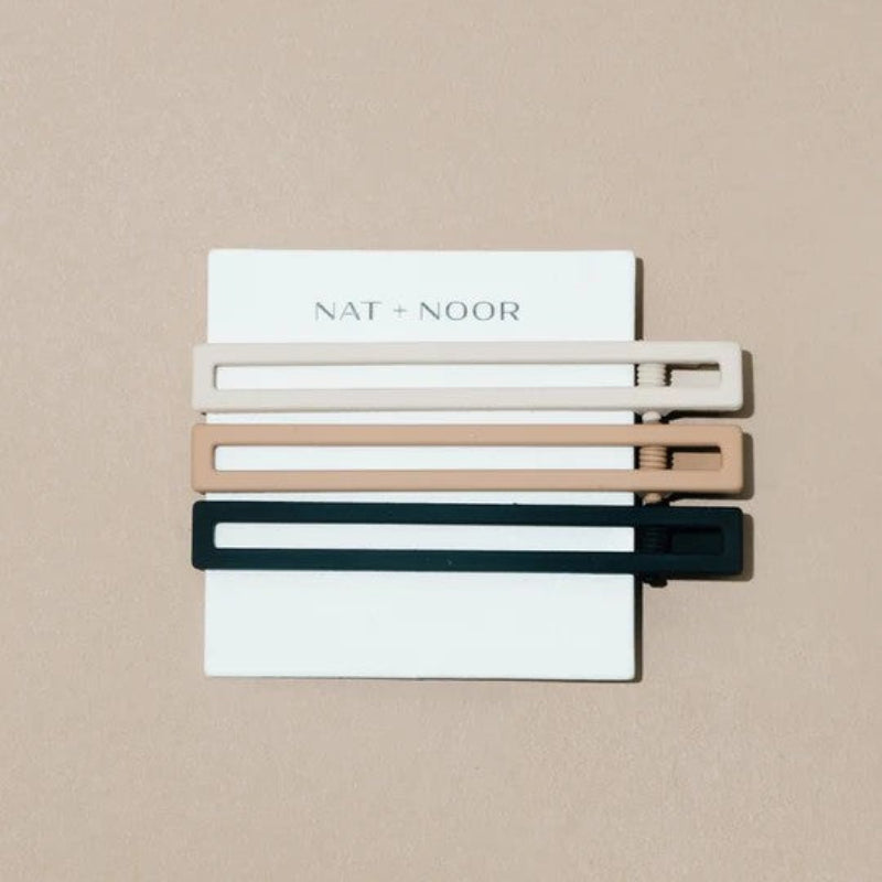 A set of three metal hair clips in white, tan, and black. The clips are attached to a white card and on a beige background. 