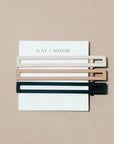 A set of three metal hair clips in white, tan, and black. The clips are attached to a white card and on a beige background. 
