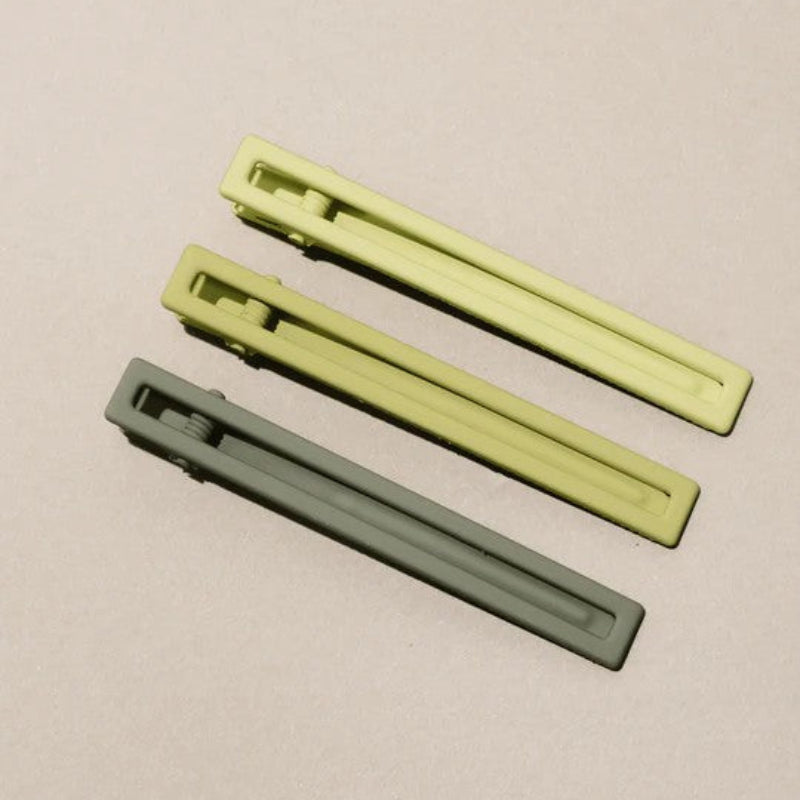 Three metal hair clips in three different shades of green on a beige background. 