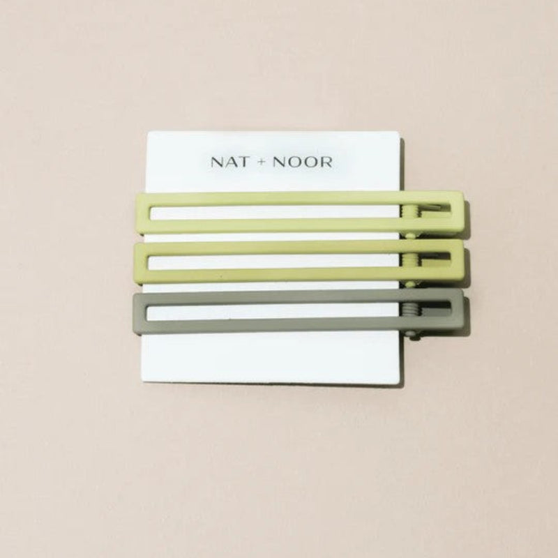 Three green metal hair clips on a white card with 'NAT + NOOR' branding against a beige background.