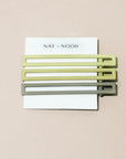 Three green metal hair clips on a white card with 'NAT + NOOR' branding against a beige background.