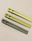 Three metal hair clips in three different shades of green on a beige background. 