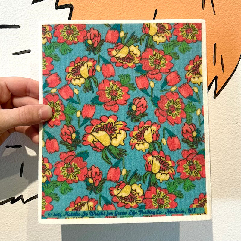 A hand holds the Potluck Press Swedish Dishcloth, a vibrant reusable cleaning cloth featuring a densely packed floral design with red, yellow, and pink flowers and green leaves on a blue background.