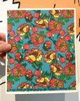 A hand holds the Potluck Press Swedish Dishcloth, a vibrant reusable cleaning cloth featuring a densely packed floral design with red, yellow, and pink flowers and green leaves on a blue background.