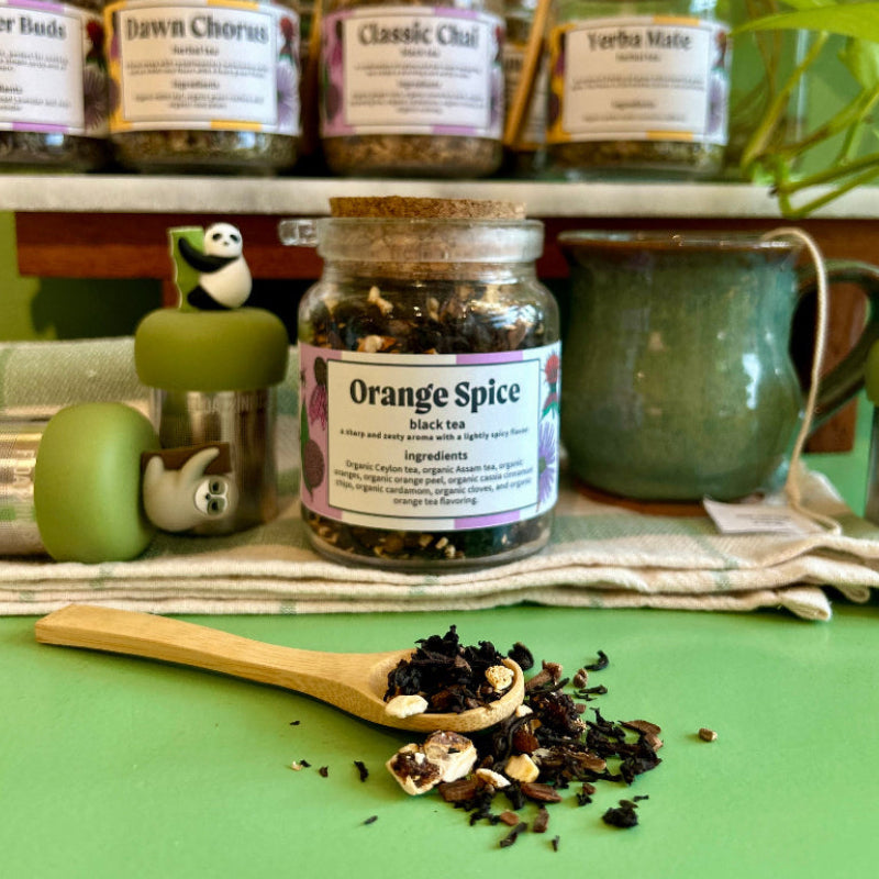 A refillable jar of orange spice loose leaf tea. The jar has a cork lid and an attachable bamboo spoon. The spoon is in front full of orange spice tea. Behind the jar is a shelf full of tea jars. 