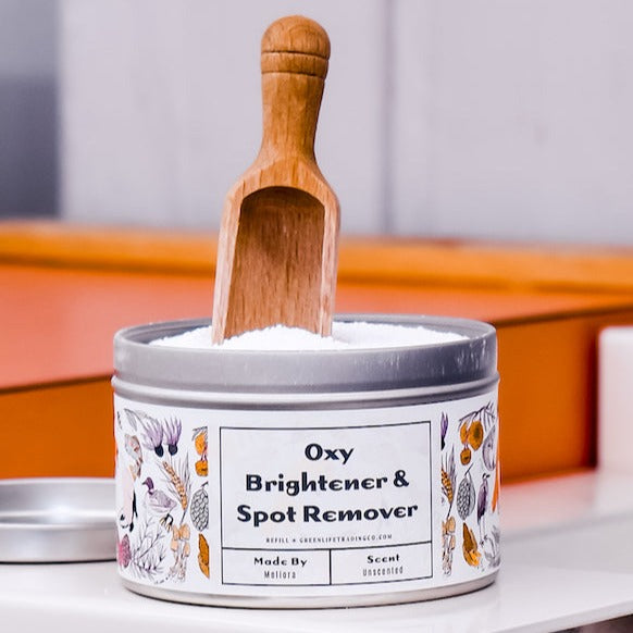 An open metal tin of Meliora Oxy Brightener & Spot Remover with a floral design displays eco-friendly white powder inside, topped with a small wooden scoop resting upright.
