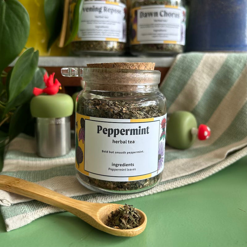 A refillable Jar of peppermint tea. The jar has a cork lid and an attachable bamboo spoon. 
